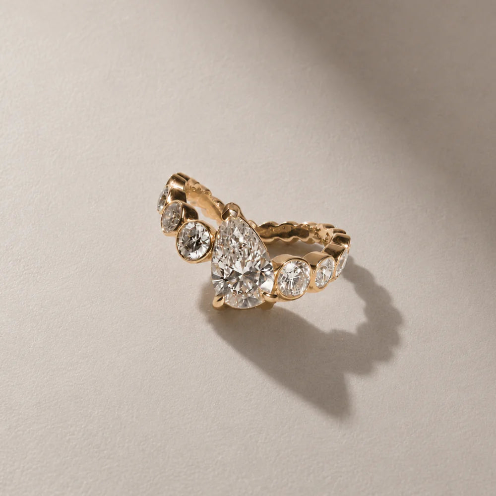 Pear diamond engagement ring with curved bezel-set round diamond band in yellow gold by ARIAH BELVI